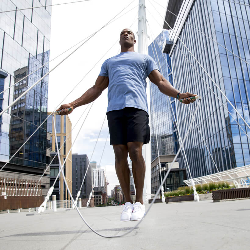 Power Starter Jump Rope Set A man jumping rope in an urban plaza between modern glass skyscrapers, wearing a light blue t-shirt, black shorts, and white sneakers.