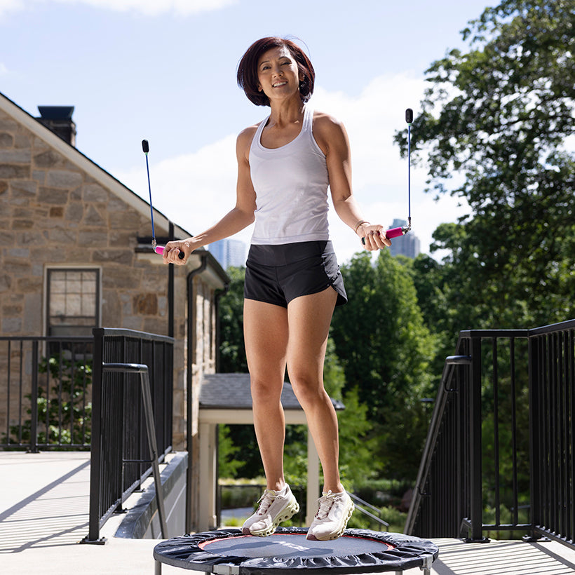 Ropeless AMP Vibe Set (LE) Woman jumping on a rebounder trampoline outdoors with Ropeless jumpe ropes with trees and a building in the background